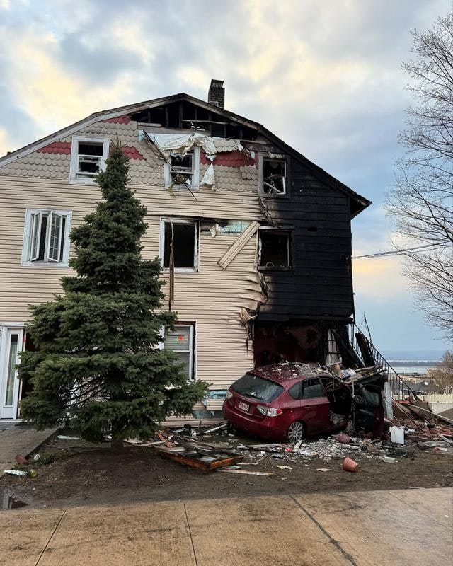 A damaged house with blackened exterior and broken windows, alongside a crumpled car and debris scattered on the ground.