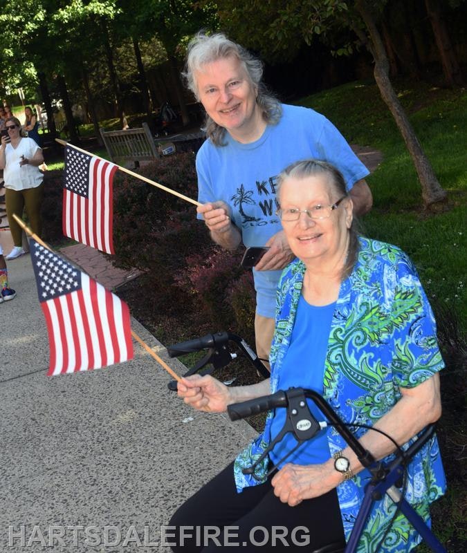 Two people with American flags, one seated with a walker, the other standing. They are outdoors on a sunny day.