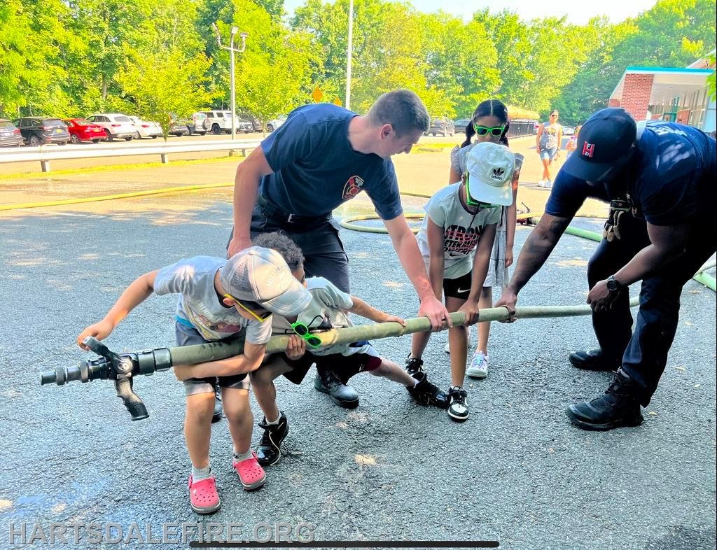 Children and firefighters handle a fire hose together outdoors.