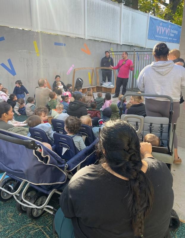 A group of children and adults watch a presentation outdoors, with some parents seated in strollers looking on attentively.