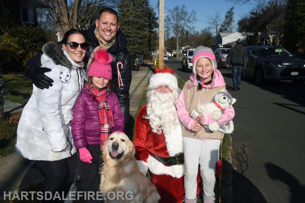 A family poses with Santa Claus and a golden retriever during a sunny winter day, enjoying a festive moment together.