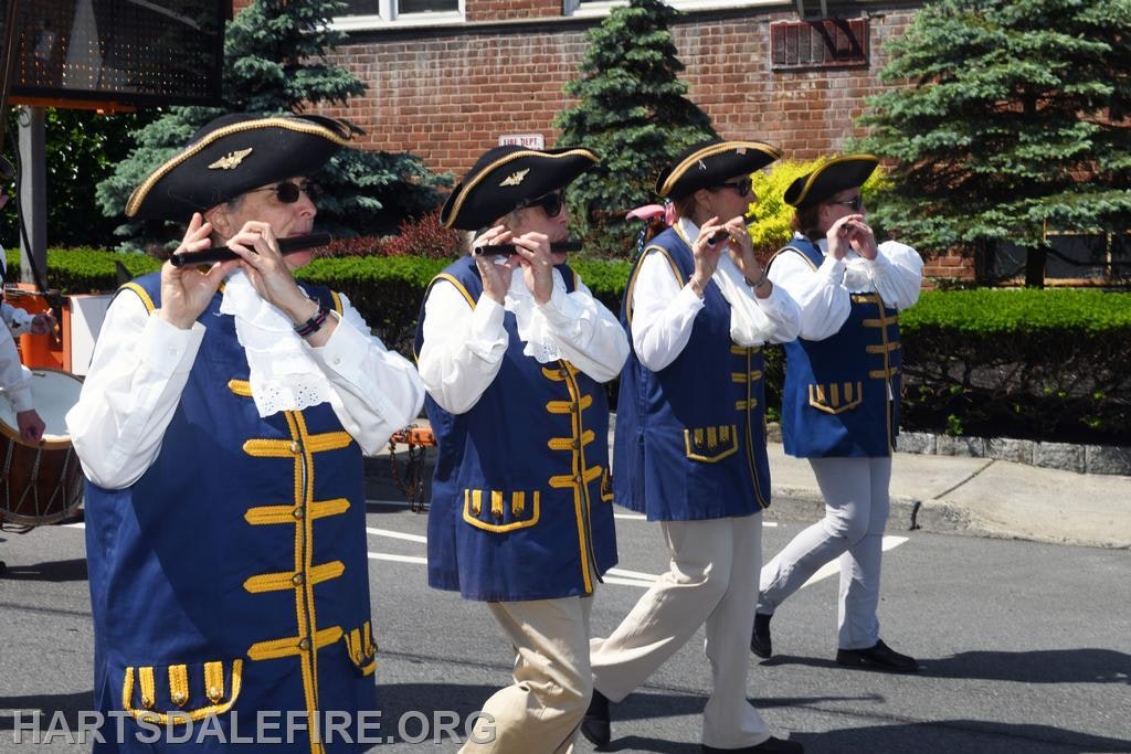 Marching musicians in colonial attire playing fifes on a sunny day.