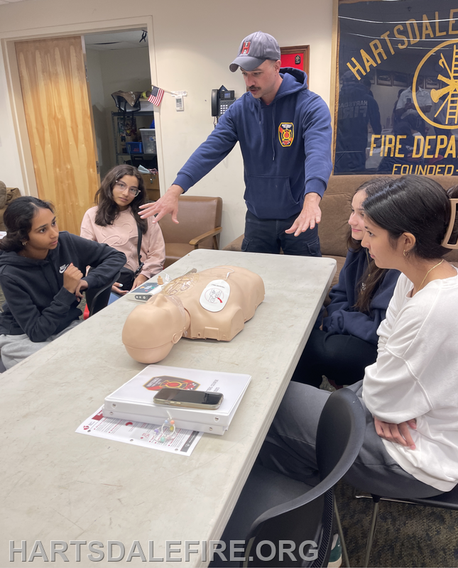 A man demonstrates CPR techniques on a training dummy to a group of interested young women in a fire department setting.