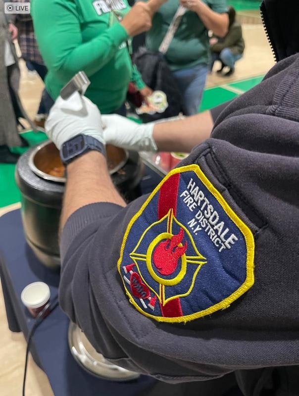 An individual in gloves is cooking or serving food, wearing a "Hartsdale Fire District" jacket, with people in the background.