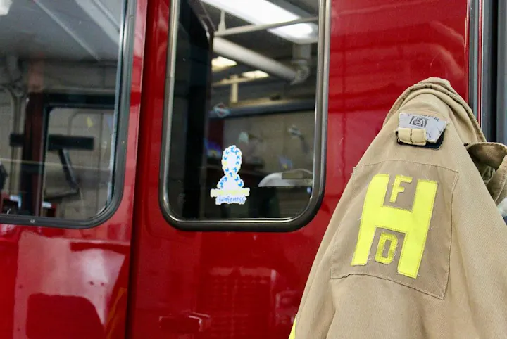 The image shows a fire station with a fire jacket hanging next to a red fire truck, featuring a sticker on the window.