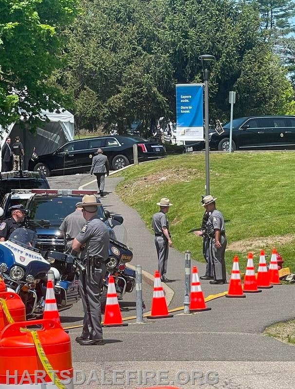 Police officers standing near motorcycles and patrol cars, with traffic cones blocking a path, and black vehicles parked in the background.