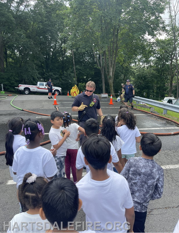 Firefighters engage with kids at an outdoor event, demonstrating fire safety with hoses while kids watch eagerly.
