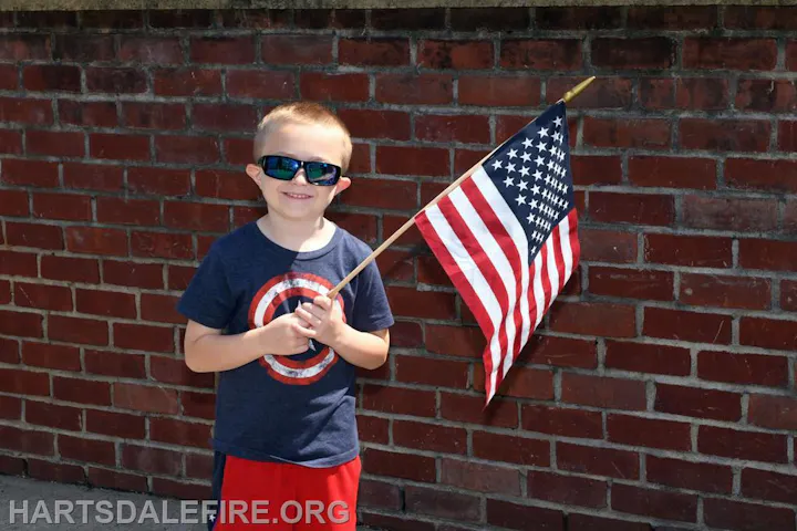 A child in sunglasses holds an American flag, standing against a brick wall.