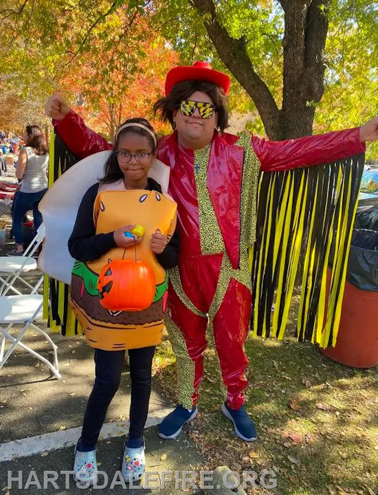 A girl in a hamburger costume poses with a man in flashy red attire, both enjoying a festive outdoor event.