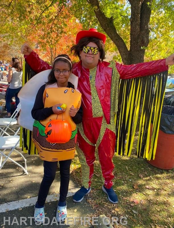 A girl in a hamburger costume poses with a man in flashy red attire, both enjoying a festive outdoor event.