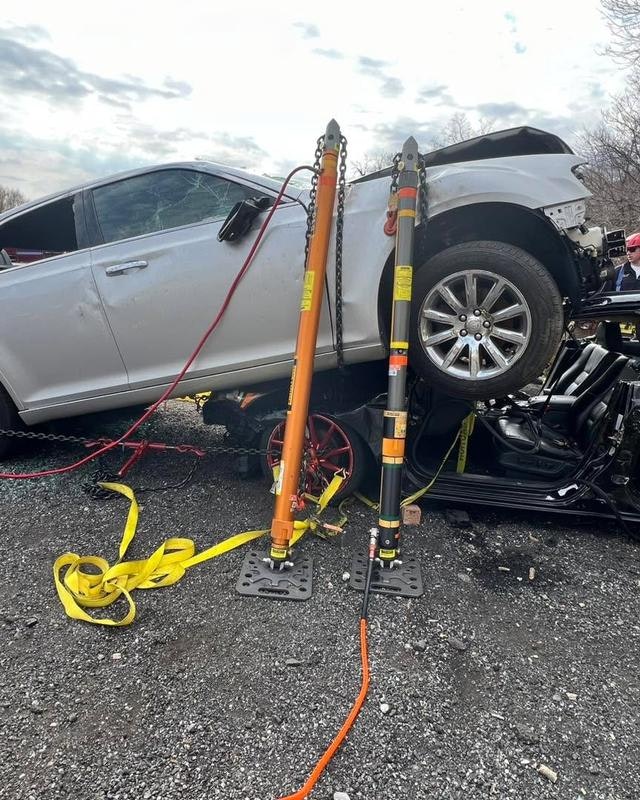 An overturned car is being stabilized and lifted using hydraulic jacks and straps in a recovery operation.