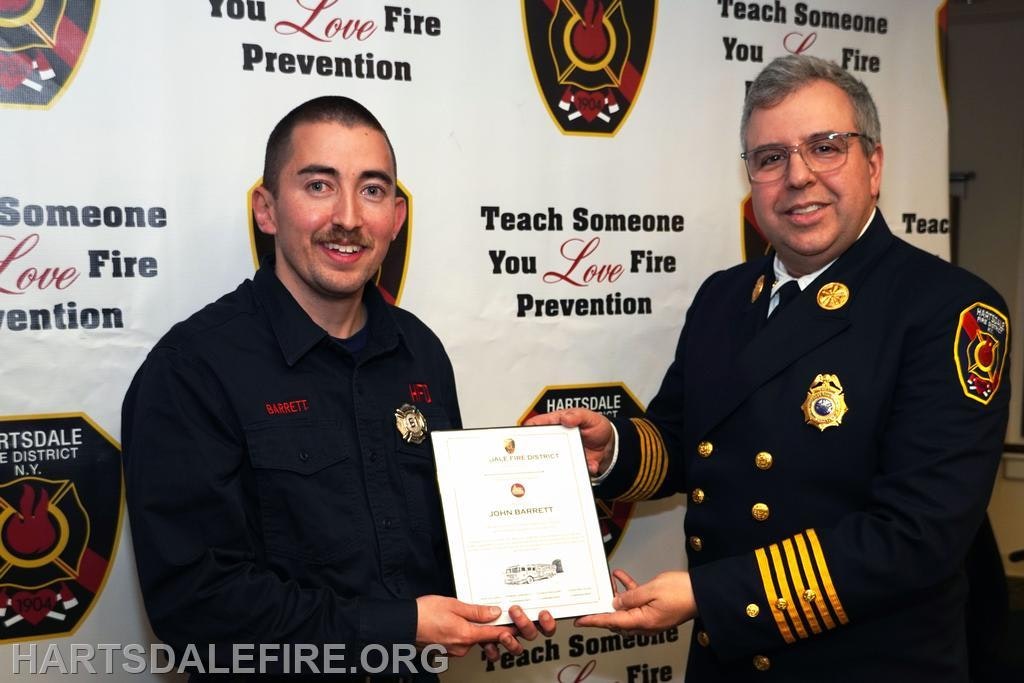 A firefighter named Barrett receives an award from a fire chief, with banners promoting fire prevention in the background.