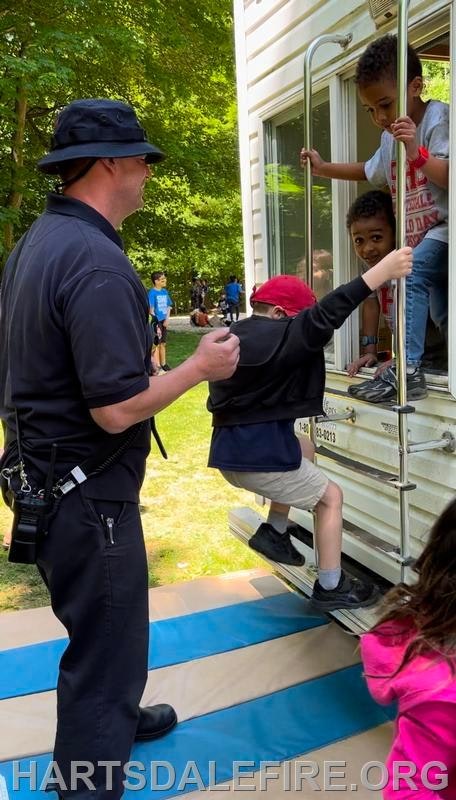 A person assists children climbing into a trailer at an outdoor event with onlookers nearby.