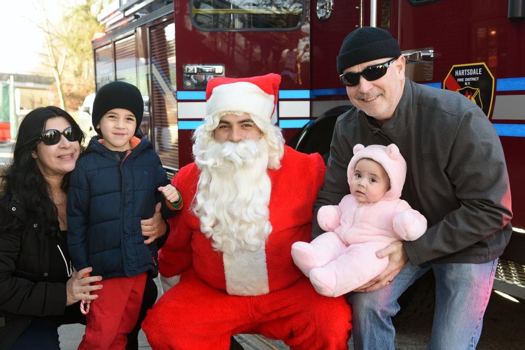 A family poses with Santa Claus in front of a fire truck, with two children dressed warmly.