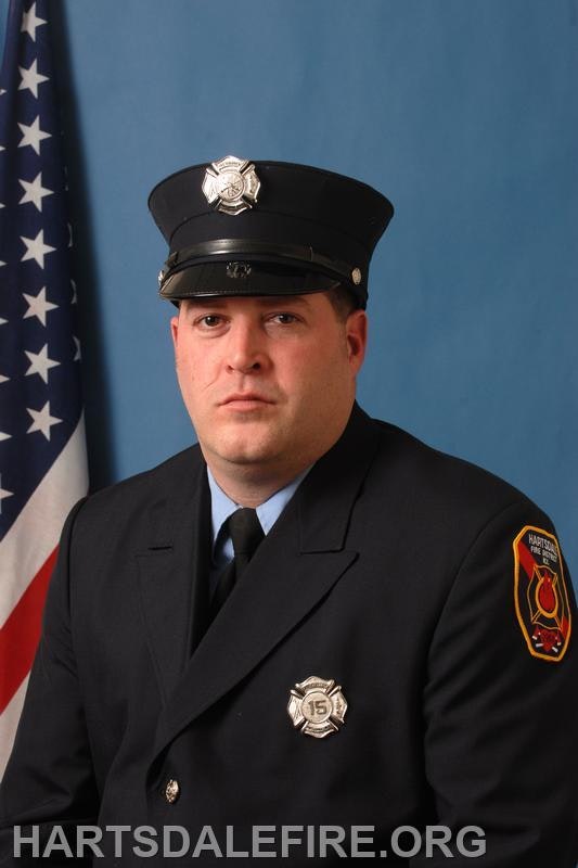 A uniformed firefighter poses against a blue background, with an American flag partially visible behind him.