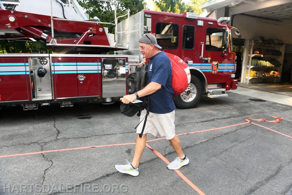 A person walks past a fire truck, wearing casual clothes and carrying a backpack, likely heading to or from a shift.