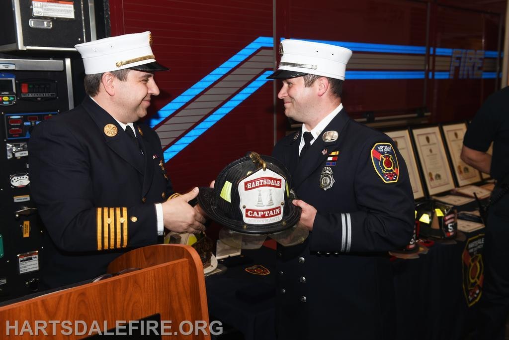 Two fire officers smiling; one hands a "Hartsdale Captain" helmet to the other.