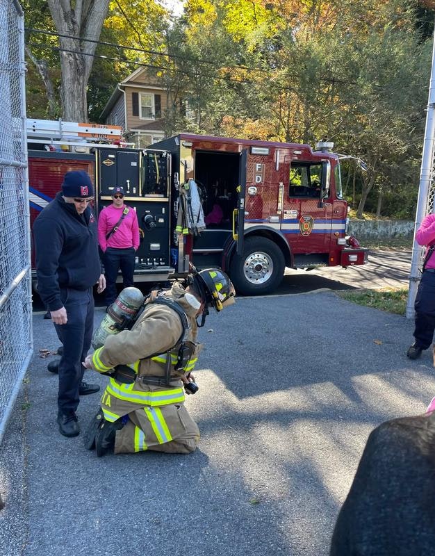 A firefighter in gear kneels near a truck while people in the background, some in pink, watch. It's a community or safety event.