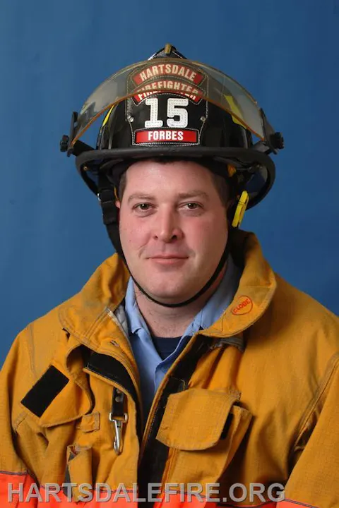A firefighter poses in uniform with a helmet displaying a badge and number, set against a blue background.