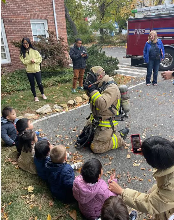 A firefighter kneels in front of a group of children, demonstrating safety, while adults watch and take photos.