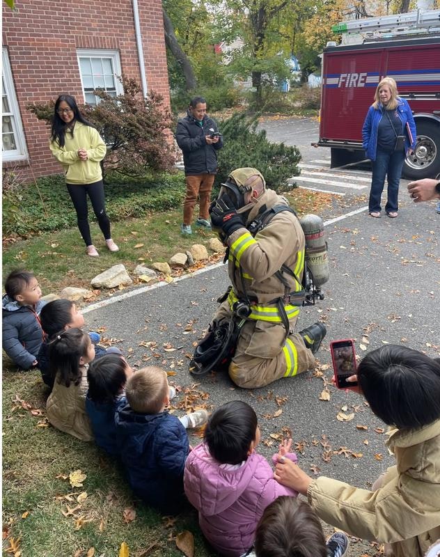 A firefighter kneels in front of a group of children, demonstrating safety, while adults watch and take photos.