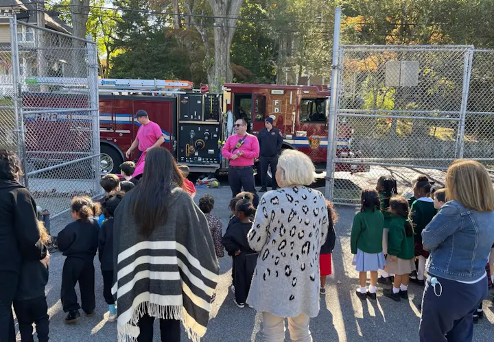 A group of children and adults are gathered near a fire truck, listening to demonstrations by firefighters in pink shirts.
