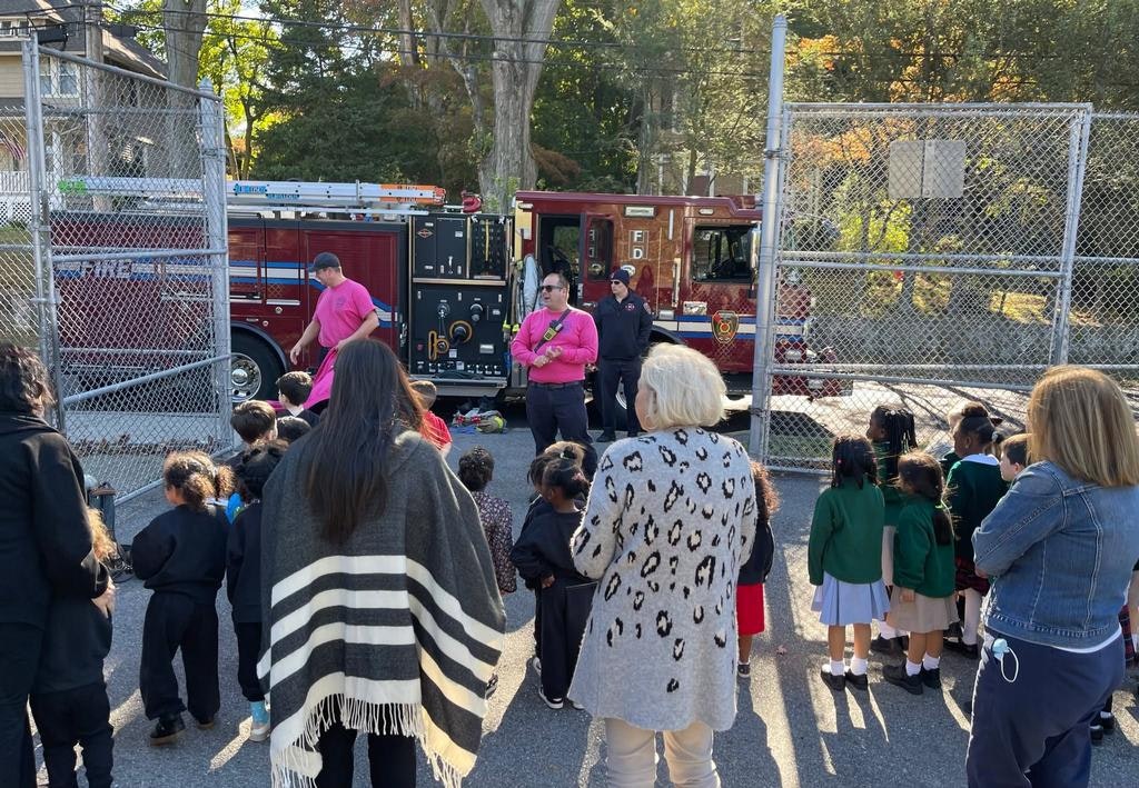 A group of children and adults are gathered near a fire truck, listening to demonstrations by firefighters in pink shirts.