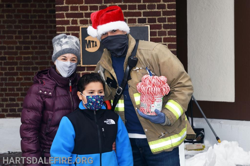 Three people in winter clothes and masks, one in a Santa hat and firefighter jacket, holding candy canes, standing in front of a brick wall.