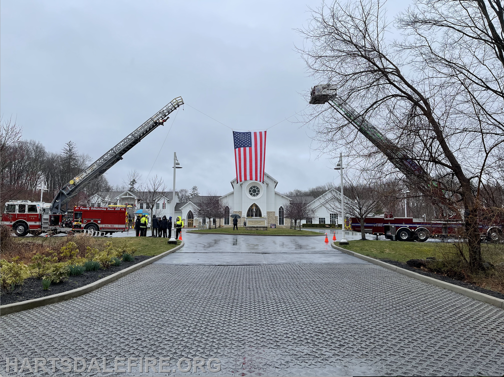 Fire trucks displaying an American flag in a rainy setting, with people gathered nearby near a church or community building.