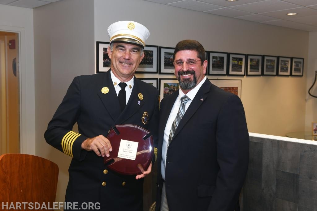 Two men smiling, one in a firefighter uniform holding a plaque, standing in an office with framed pictures on the wall.