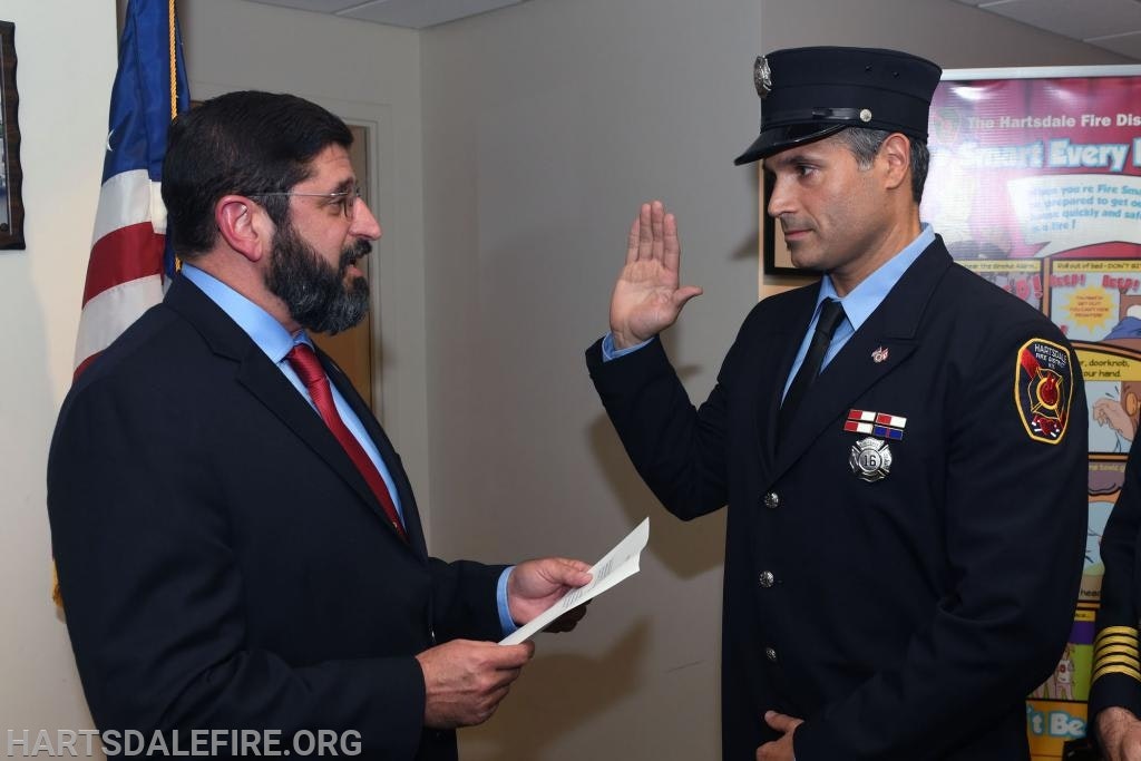 A firefighter in uniform is taking an oath with a suited man reading a document, standing next to an American flag.