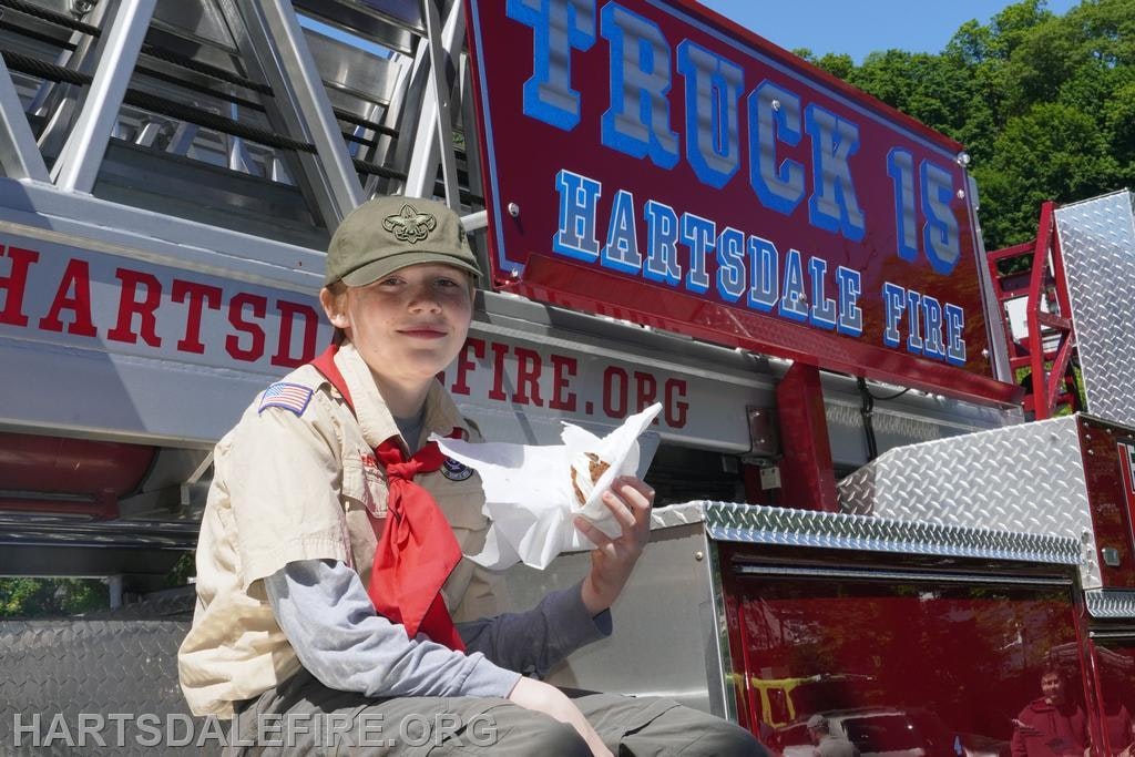 A young person in a scout uniform enjoys food next to a fire truck labeled "Hartsdale Fire." Bright day and green trees in background.