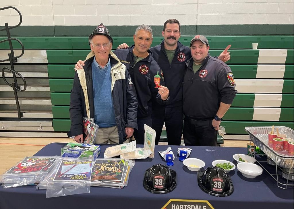 A group photo of four people, including firefighters, at a table with food and community materials, showcasing teamwork and safety.