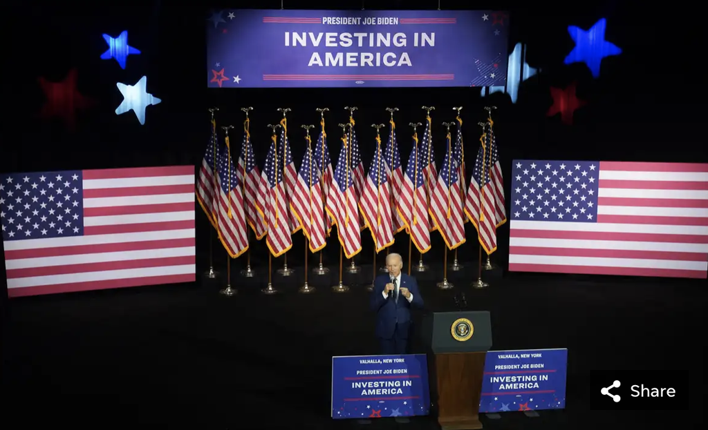 A person speaking at a podium with "Investing in America" signage, surrounded by U.S. flags and decorations.