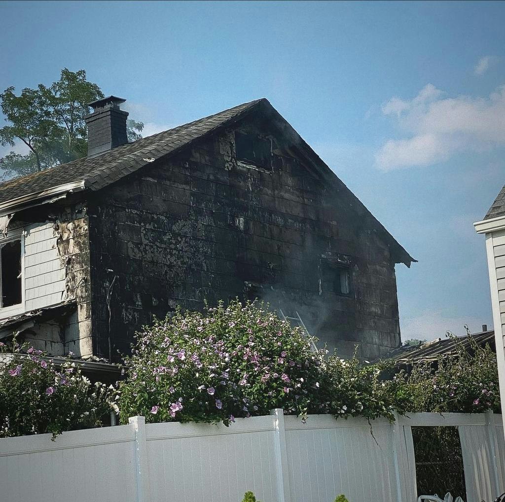 A partially burned house showing damage with smoke rising, surrounded by blooming flowers and a white fence, under a clear sky.