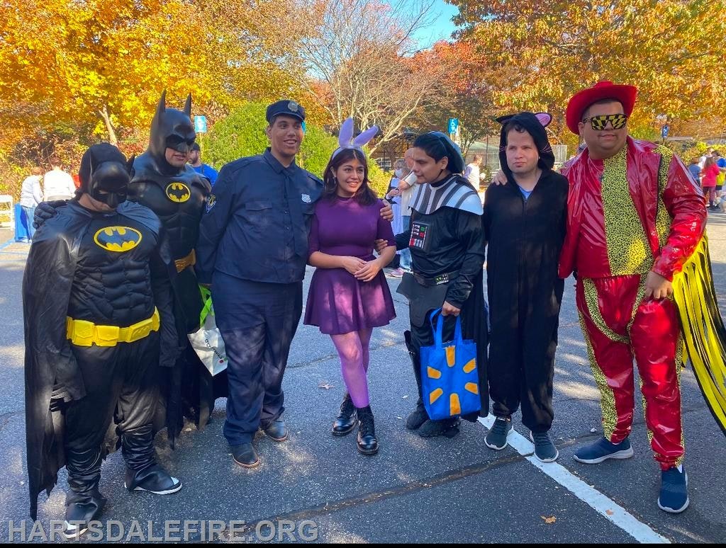 A group of people in various costumes, including superheroes and characters, posing outdoors with colorful autumn trees in the background.