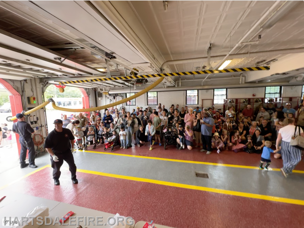 A large crowd gathers inside a fire station, with firefighters present and people taking photos, enjoying a community event.