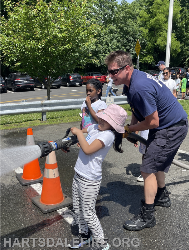 A firefighter helps a child operate a fire hose, while another girl watches, at a community event outdoors.