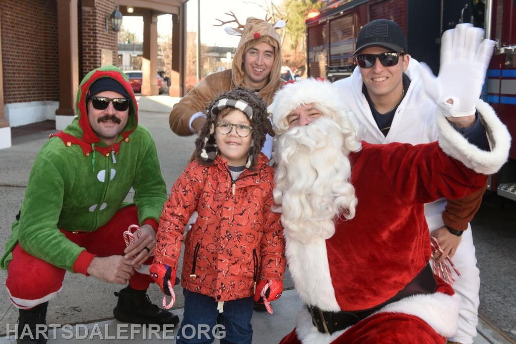 A group of costumed individuals, including Santa, pose with a child, spreading holiday cheer.