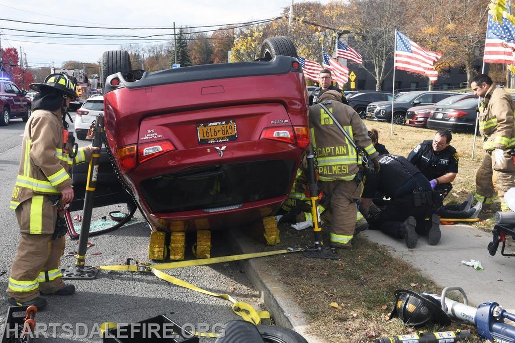 Emergency responders assist at the scene of a flipped over car, with firefighters and police providing aid. Flags are visible nearby.