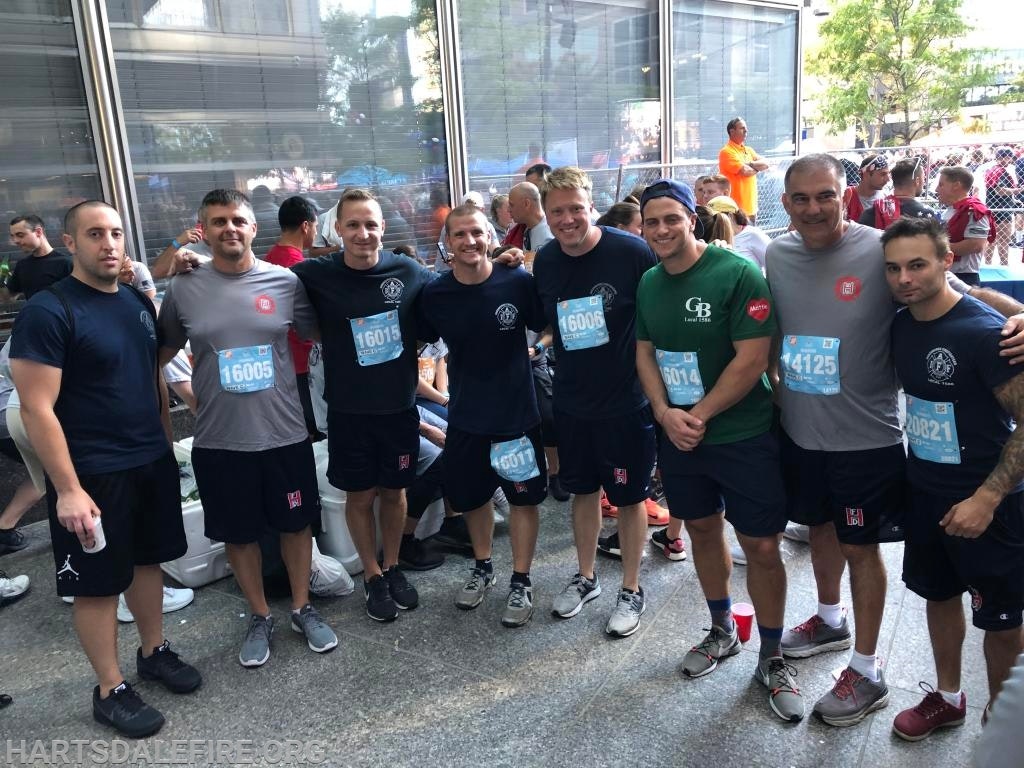 A group of eight men in athletic gear, wearing numbered race bibs, pose together outside a building.