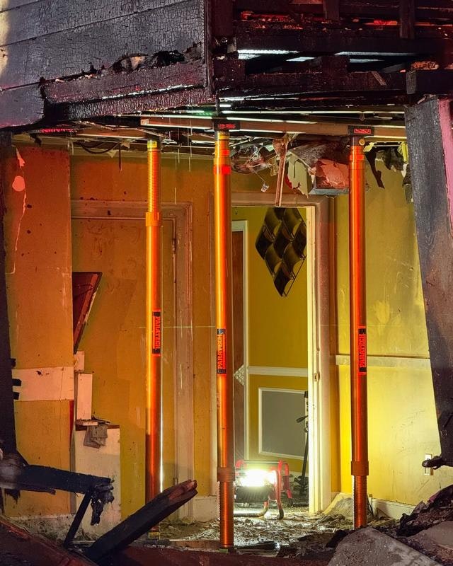 The image shows a damaged interior with charred walls, support props in place, and a faint light illuminating the space.