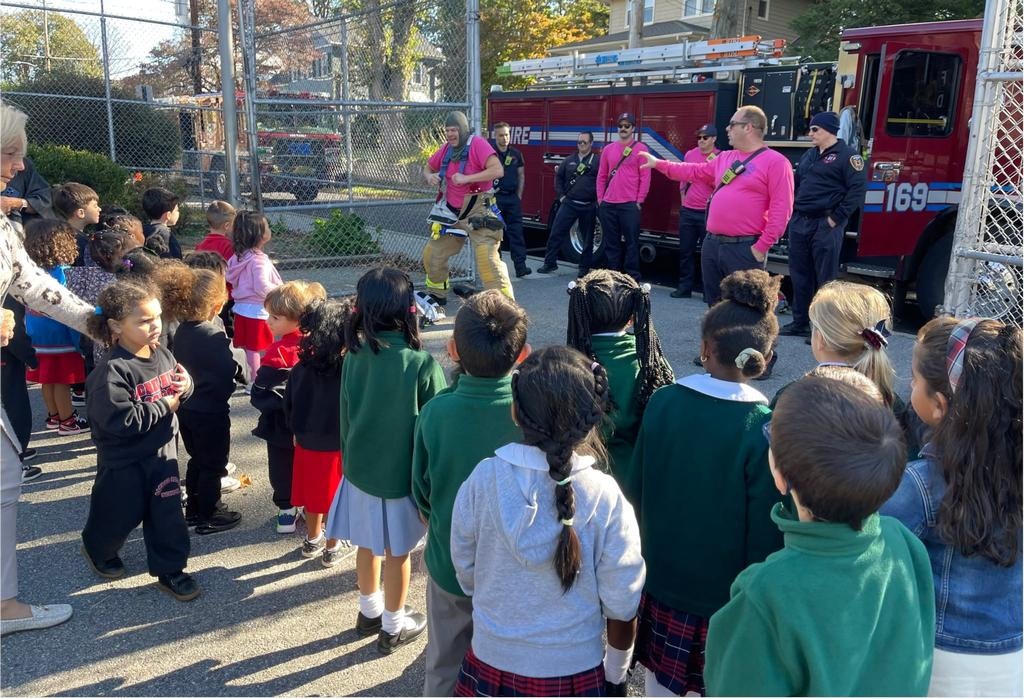 A group of children watches firefighters demonstrate safety procedures near a fire truck, with adults supervising the event.