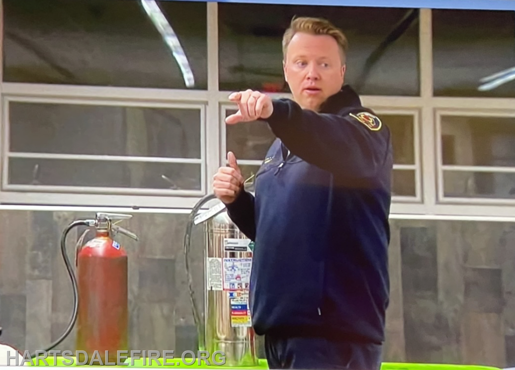A person in a uniform points while standing near fire safety equipment, suggesting a demonstration or instruction.