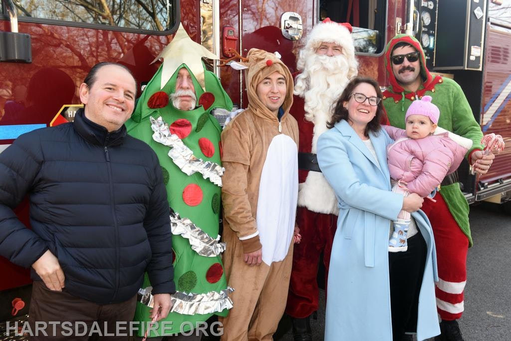 A festive group poses with Santa and holiday characters near a fire truck, capturing a cheerful holiday spirit.