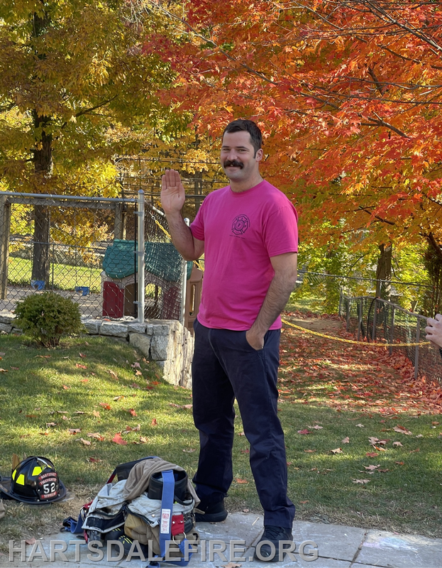 A man in a pink shirt and work pants stands outdoors surrounded by autumn foliage, raising his hand in a friendly gesture.