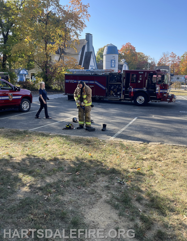 A firefighter prepares gear in a parking lot near a fire truck, while a woman walks nearby on a sunny day.