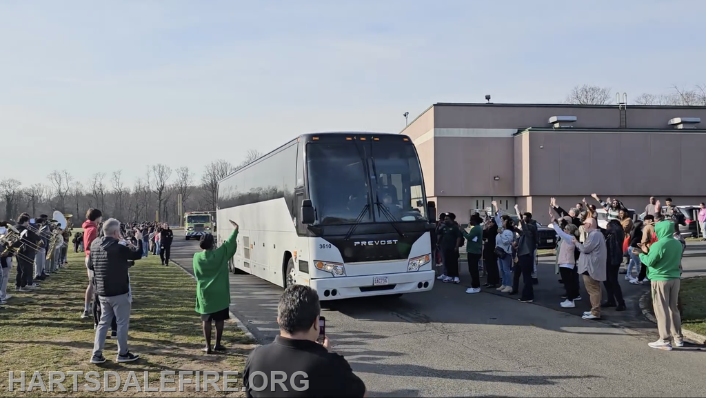 A crowd waves and cheers as a large bus drives by, with a band performing nearby, in a celebratory outdoor setting.
