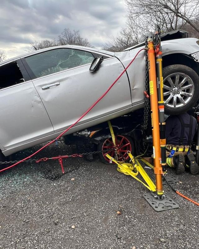 A silver car is tilted at an angle, secured with chains and tools, likely after an accident, with responders working nearby.