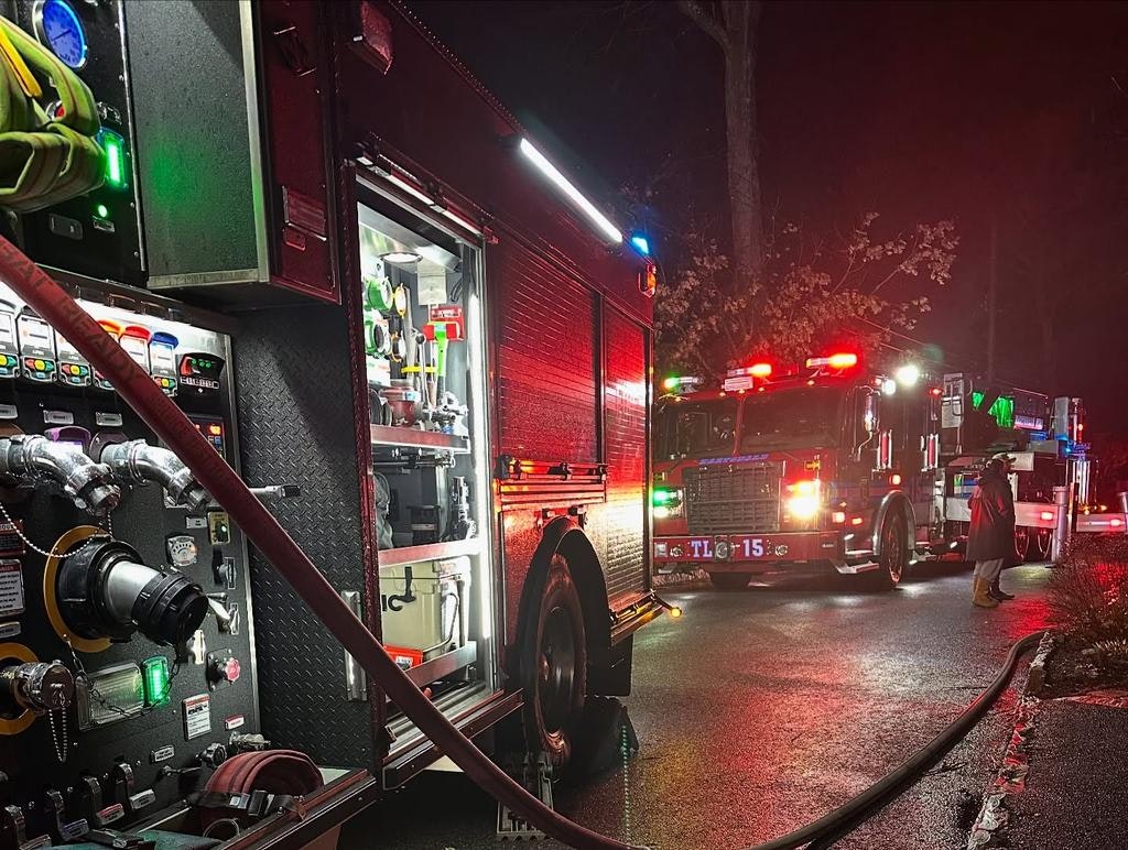 A scene at night featuring fire trucks and equipment, with flashing lights and hoses visible, indicating a response to an emergency.